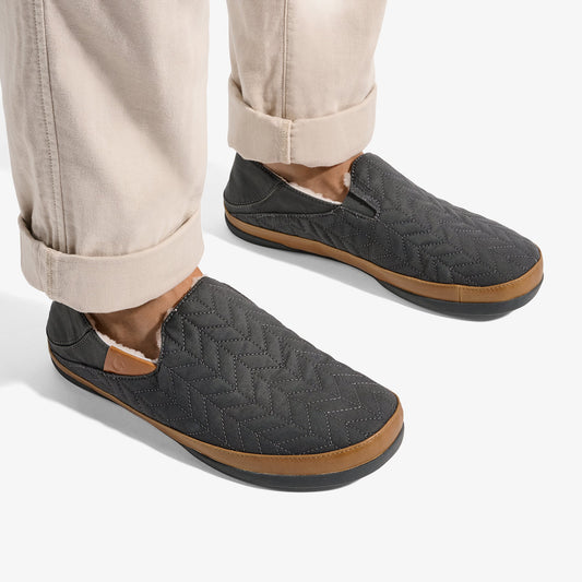 Dark gray quilted slip-on slippers with tan rubber soles, worn with rolled cream pants.